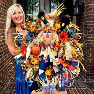 May include: A large, colorful scarecrow wreath with a straw-filled head, a plaid shirt, and blue overalls. The wreath is decorated with sunflowers, pumpkins, and ribbons, creating a festive autumn theme.