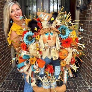 May include: A decorative scarecrow wreath featuring a central scarecrow figure with a tan hat, straw hair, and a tag reading "Country Crow". The wreath is adorned with teal and orange sunflowers, small pumpkins, ribbons, and black bird accents.