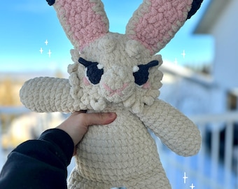 May include: A crocheted plush bunny toy with pink ears, black accents, and a white body. The bunny has a grumpy expression with black eyes and a small black nose.