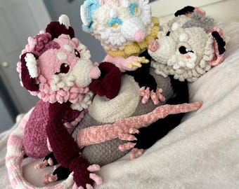 May include: Three crocheted stuffed animals, a pink and white opossum, a white and blue opossum, and a gray and white opossum, are lying on a white blanket.