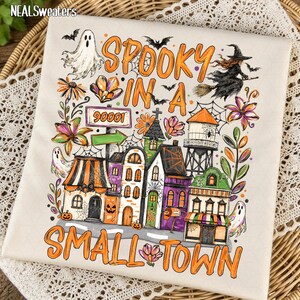 Puede incluir: Una tela de color crema con un gráfico colorido de temática de Halloween con el texto "SPOOKY IN A SMALL TOWN" y "90001". El diseño incluye una bruja en una escoba, fantasmas, murciélagos, una torre de agua y varios edificios con detalles en naranja.
