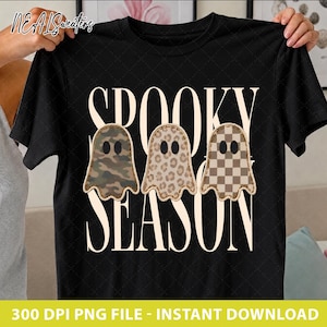 Op de afbeelding: Een zwart t-shirt met korte mouwen en de tekst SPOOKY SEASON in grote crèmekleurige letters. Drie spookillustraties staan in het midden van het shirt, elk met een ander patroon: groen camouflage, bruine luipaardprint en een bruin dambordpatroon.