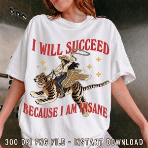 May include: A white t-shirt featuring a graphic of a cowgirl riding a winged tiger while holding a lasso. The text "I WILL SUCCEED" is printed in red above the graphic, and "BECAUSE I AM INSANE" is printed in red below the graphic.