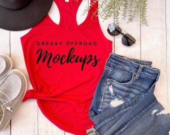 May include: Red racerback tank top with the text "Greasy Offroad Mockups" printed on the front. The tank top is laying on a white wooden surface with a pair of jeans, a hat, and sunglasses.