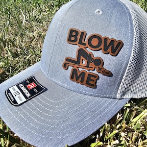 May include: A pale blue trucker cap with a white mesh back. The hat displays a brown leather patch with the words "BLOW ME" and a graphic illustration. It has an adjustable strap and a Richardson brand tag.