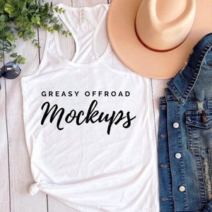 Puede incluir: Camiseta sin mangas blanca con el texto "GREASY OFFROAD MOCKUPS" en negro. La imagen también incluye un sombrero beige, una chaqueta vaquera, gafas de sol y vegetación, todo dispuesto sobre una superficie de madera clara.