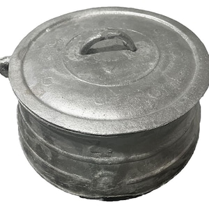 May include: A round, silver-colored metal pot with a matching lid and two side handles. The pot features a textured, cast-metal surface with horizontal ridges around the body and a central handle on the circular lid, showing a sturdy construction.
