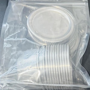 May include: A stack of circular, silver-colored metal lids enclosed within a clear, resealable plastic storage bag. The lids feature a smooth, flat top surface with a slightly raised, rounded rim, designed for sealing containers securely.