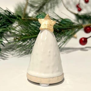 May include: A conical ceramic tree figurine with a glossy white textured surface and a matte beige base. The top of the tree is adorned with a five-pointed star finished in a metallic gold glaze, creating a distinct contrast against the white body.