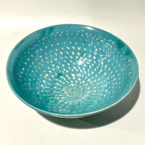 May include: A turquoise ceramic bowl with a spiral pattern of small, white, triangular cutouts. The bowl has a smooth, glossy finish and a rounded shape. The bowl is a decorative piece.