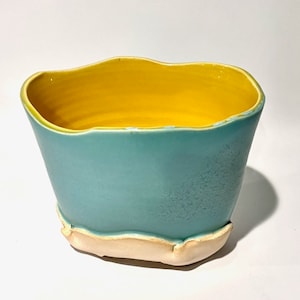 May include: A ceramic planter with a light blue glaze and a yellow rim. The planter has an irregular, wavy shape and a white base.