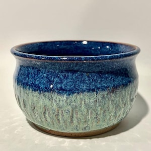 May include: A handcrafted ceramic bowl with a textured surface. The bowl features a gradient of colors, with a deep blue rim transitioning to a lighter blue and green lower section. The base is a natural brown color.