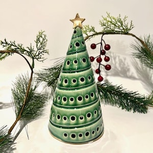 May include: A conical ceramic Christmas tree sculpture in a glossy green glaze with horizontal ridges and circular cutouts. The tree is topped with a small, five-pointed gold star. The surface features a gradient of light to dark green.