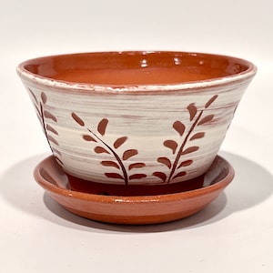 May include: Terracotta planter with a white glaze and brown leaf design. The planter has a matching saucer.