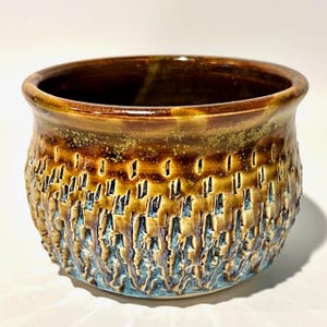 May include: A handcrafted ceramic bowl with a textured exterior and a glossy, glazed finish. The bowl features a blend of brown, gold, and blue hues. The interior is a dark brown color. This decorative bowl is suitable for home decor.