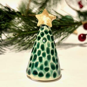 May include: A cone-shaped ceramic Christmas tree figurine with a glossy light green glaze and dark green circular spots. The tree is topped with a textured, gold-colored star ornament. The surface of the ceramic has a dimpled, uneven texture.