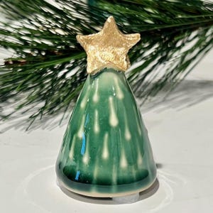 May include: A conical ceramic tree figurine with a glossy dark green glaze and white drip-like accents. The tree is topped with a textured, metallic gold-colored star. The surface of the ceramic is smooth and reflective throughout the piece.