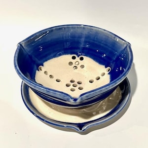 May include: A handmade ceramic colander and matching saucer. The colander is a deep blue with a cream-colored interior and drainage holes. The saucer is cream-colored with a blue rim. The set is ideal for kitchen use.