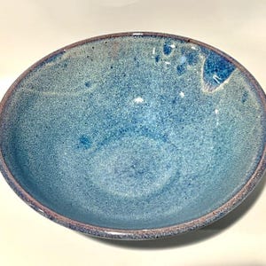 May include: A handmade ceramic bowl with a speckled blue glaze. The bowl has a rounded shape with a slightly raised rim. The rim is a darker shade of blue, and the interior has a mottled appearance.