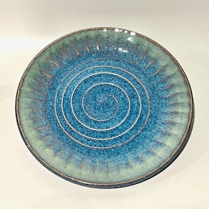 May include: A handmade ceramic plate with a spiral design. The plate features a gradient of blue and green hues, with a dark rim and a series of concentric white lines at the center. The plate has a diameter of approximately 8 inches.