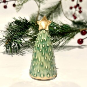 May include: A cone-shaped ceramic Christmas tree figurine in a glossy sage green glaze with raised white drip-like accents. The tree is topped with a textured, metallic gold star. The base is a smooth, unglazed cream-colored ceramic.