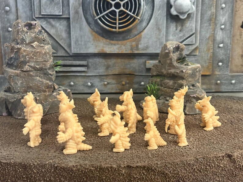 Peut inclure: Une collection de onze petites figurines de monstres en plastique beige clair non peintes. Chaque cr&eacute;ature pr&eacute;sente une posture bip&egrave;de, une peau textur&eacute;e avec des pointes et des traits distincts. Les figurines sont dispos&eacute;es en groupe sur une surface textur&eacute;e.