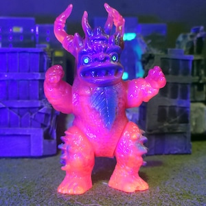 May include: A neon pink and orange monster figurine with blue detailing. The toy features horns, a toothy grin, and glowing blue eyes. It is posed with its arms raised, set against a miniature cityscape.