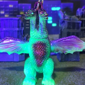 May include: A two-headed dragon figurine with green and purple detailing. The dragon has two heads, wings, and a textured body. The figurine is set against a backdrop of miniature buildings, with a neon green glow.