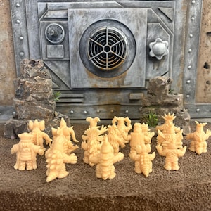 Peut inclure: Une collection de petites figurines de monstres couleur p&ecirc;che clair pos&eacute;es sur une surface brune textur&eacute;e devant une structure murale grise de style industriel avec une grille circulaire et des d&eacute;tails m&eacute;talliques. Les figurines pr&eacute;sentent divers designs.