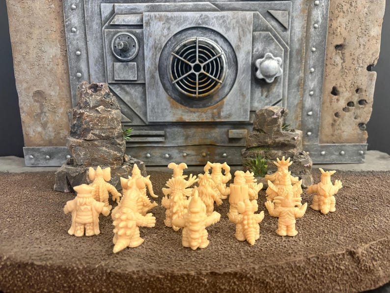 Peut inclure: Une collection de petites figurines de monstres couleur p&ecirc;che clair pos&eacute;es sur une surface brune textur&eacute;e devant une structure murale grise de style industriel avec une grille circulaire et des d&eacute;tails m&eacute;talliques. Les figurines pr&eacute;sentent divers designs.
