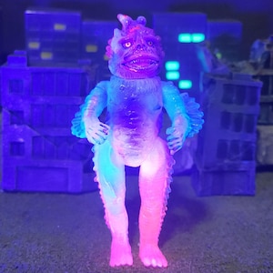 May include: A brightly coloured monster figurine with a detailed face and textured body. The figure is mainly pink and blue, with a purple head and is standing in front of a cityscape. The lighting gives the figure a neon appearance.