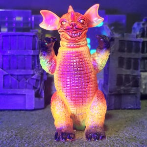 May include: A brightly coloured monster figurine in shades of orange and yellow, with a textured body, large ears, and a toothy grin. The toy has black claws and raised arms. The background shows miniature city buildings.
