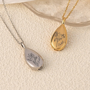 May include: Two teardrop-shaped pendant necklaces. One is silver-toned, the other gold-toned. Each pendant features a floral design and a name engraved on the front. The necklaces are displayed on a light-colored surface.