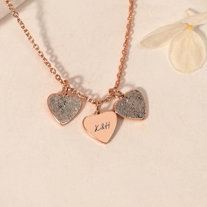 Può includere: Collana in oro rosa con tre ciondoli a forma di cuore. Due cuori presentano disegni di impronte digitali e il cuore centrale ha le iniziali "X&H" incise. La collana è esposta su una superficie bianca con un fiore.