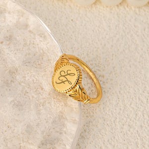 Könnte beinhalten: Ein goldfarbener Siegelring mit einem eingravierten Buchstaben "L" auf einer ovalen Fläche. Der Ring hat ein dekoratives Blattdesign am Band. Eine Perlenkette befindet sich im Hintergrund.