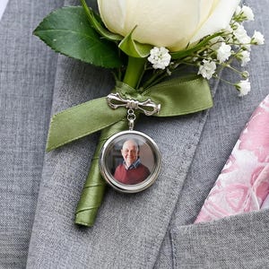 Könnte beinhalten: Grauer Anzug mit weißer Rosenknopflochblume, grünem Band und kleinem Fotohänger. Ein Einstecktuch mit rosa Blumenmuster und weißem Stoff befindet sich in der Jackentasche.