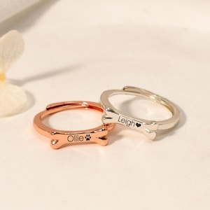 Könnte beinhalten: Zwei personalisierte Hunde-Knochenringe. Der Ring links ist roségoldfarben und trägt die Aufschrift "Ollie" mit einem Pfotenabdruck. Der Ring rechts ist silberfarben und trägt die Aufschrift "Leigh" mit einem Herz.