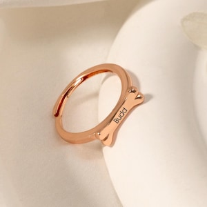 Könnte beinhalten: Roségoldener verstellbarer Ring in Knochenform mit der Gravur "Buda".