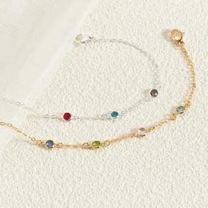 以下が含まれることがあります： 小さなカラフルな宝石が付いた繊細なブレスレットが2つ。上のブレスレットはシルバーで、赤、青、紫の宝石が付いています。下のブレスレットはゴールドで、青、緑、透明な宝石が付いています。