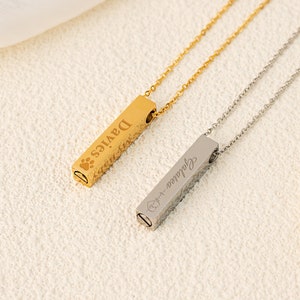 Può includere: Due collane a barra d'oro e d'argento con nomi incisi, "DAVIES" e "Julianna", su sfondo bianco. Le collane sono entrambe su una superficie bianca con una collana di perle.