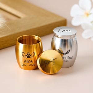 Könnte beinhalten: Zwei kleine Urnen aus Metall, eine goldfarbene und eine silberfarbene. Die goldene Urne zeigt einen Pfotenabdruck mit Engelsflügeln und den Text Alice 2021-2026. Der Deckel ist mit Love You graviert. Die silberne Urne trägt den Namen Emma und einen Zweig.