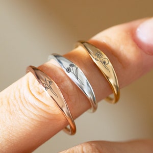 Könnte beinhalten: Drei Silber- und Goldringe mit einem zarten, gravierten Blumenmuster auf jedem Ring. Die Ringe sind auf einem Finger übereinander gelegt.