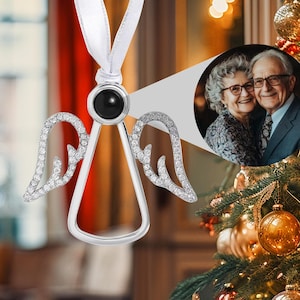 May include: Silver angel Christmas tree ornament with crystal wings and a black stone. It hangs from a white ribbon. A small photo of a couple is in the background, near a decorated Christmas tree.