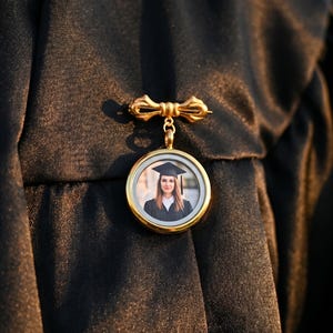 May include: A gold graduation pin featuring a bow design and a circular pendant. The pendant displays a photograph of a person in a graduation cap and gown. The pin is fastened to a black graduation gown.