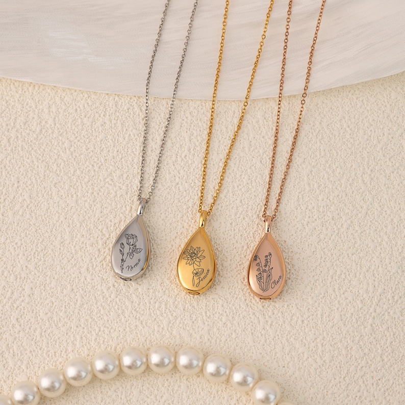 May include: Three teardrop-shaped necklaces in silver, gold, and rose gold. Each pendant features a floral design and a name. The necklaces are displayed on a cream-colored surface with a pearl necklace in the foreground.