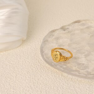 May include: A gold signet ring with an oval-shaped top featuring engraved initials. The ring has a detailed design with a textured band.