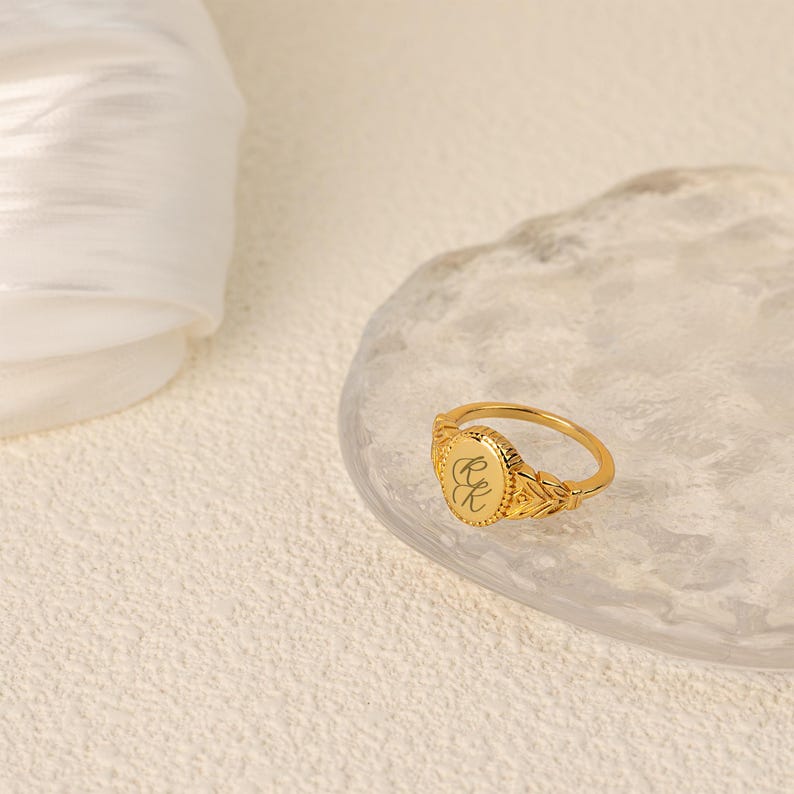 May include: A gold signet ring with an oval-shaped top featuring engraved initials. The ring has a detailed design with a textured band.