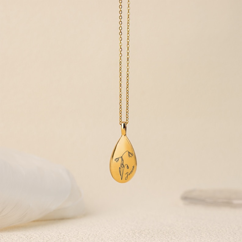 May include: A gold-colored pendant necklace with a teardrop-shaped charm. The charm features an engraved floral design and the name "Jessica" in script. The necklace chain is delicate and also gold-colored.