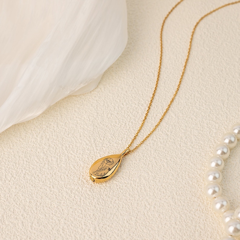 May include: A gold-colored teardrop-shaped pendant necklace with a delicate floral engraving. The pendant hangs from a fine gold chain. A strand of white pearls is visible in the background. The necklace is displayed on a textured, off-white surface.
