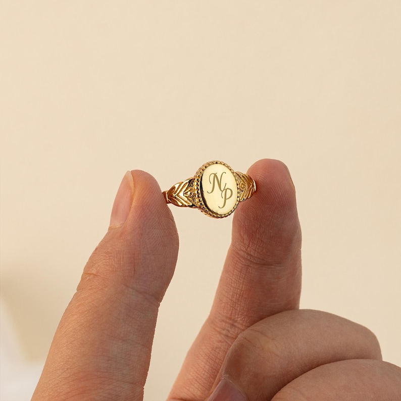 May include: Gold signet ring with a raised oval top engraved with the initials "NP". The ring has a textured band.